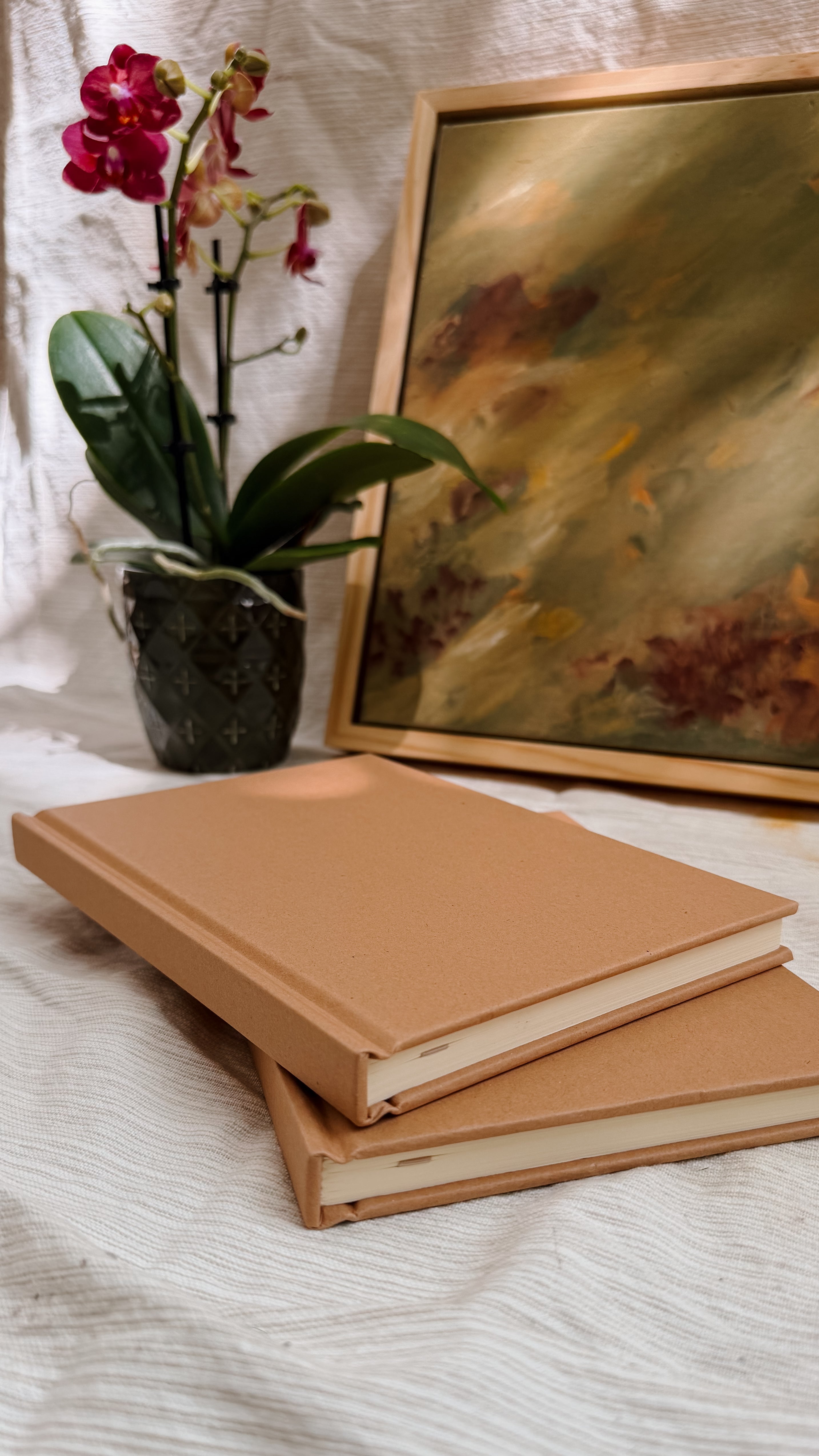 Stack of brown books with a vase of flowers and a painting in the background on a textured surface.